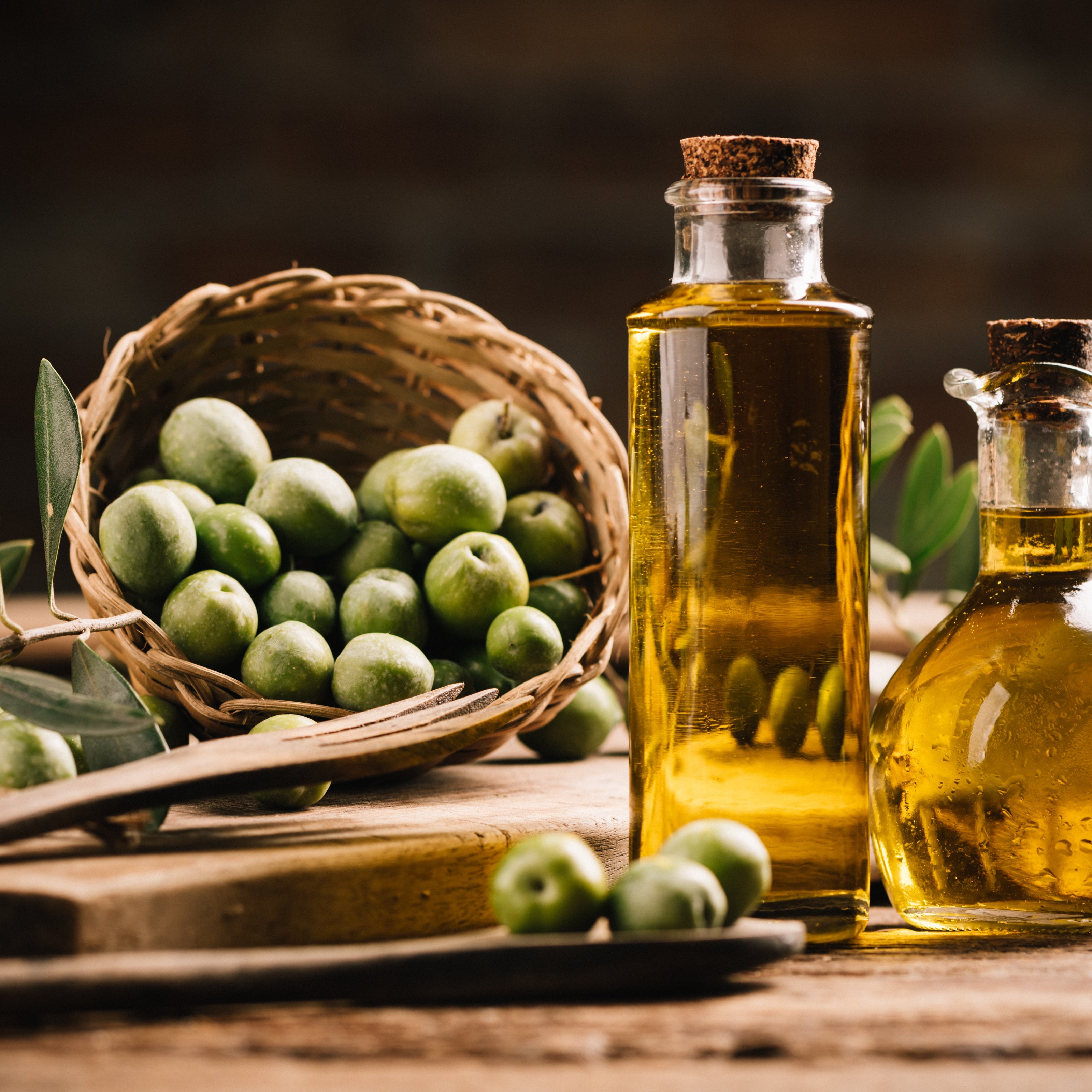 A rustic scene featuring a woven basket filled with green olives alongside two glass bottles of golden olive oil. The bottles are sealed with cork stoppers and surrounded by wooden utensils, all set against a warm, earthy background.