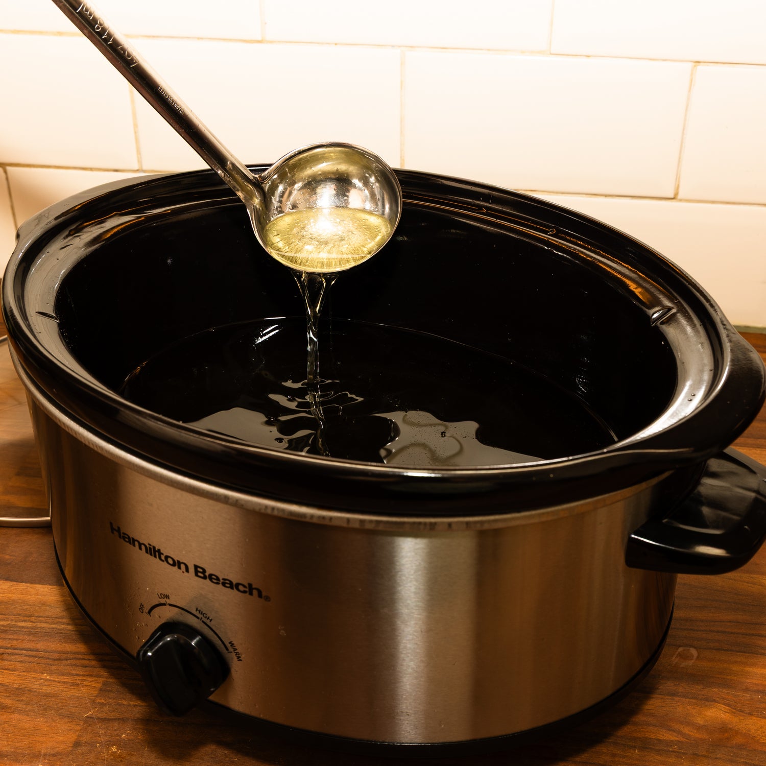 A ladle is being used to scoop clear, golden rendered tallow from a black ceramic slow cooker labeled "Hamilton Beach." The liquid appears clean and glossy, indicating it has been fully melted and strained, ready for use in balm-making or skincare formulations.