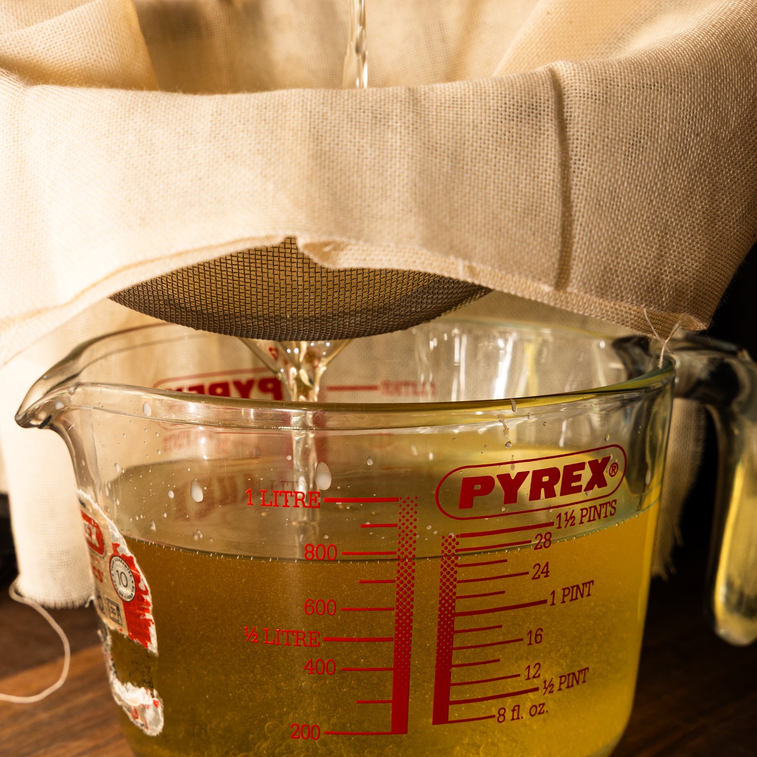 A Pyrex measuring jug is shown collecting freshly rendered liquid tallow being strained through a mesh sieve lined with cream-colored cheesecloth. The golden liquid is mid-pour, and the warm lighting highlights its clarity and purity during the filtering process.