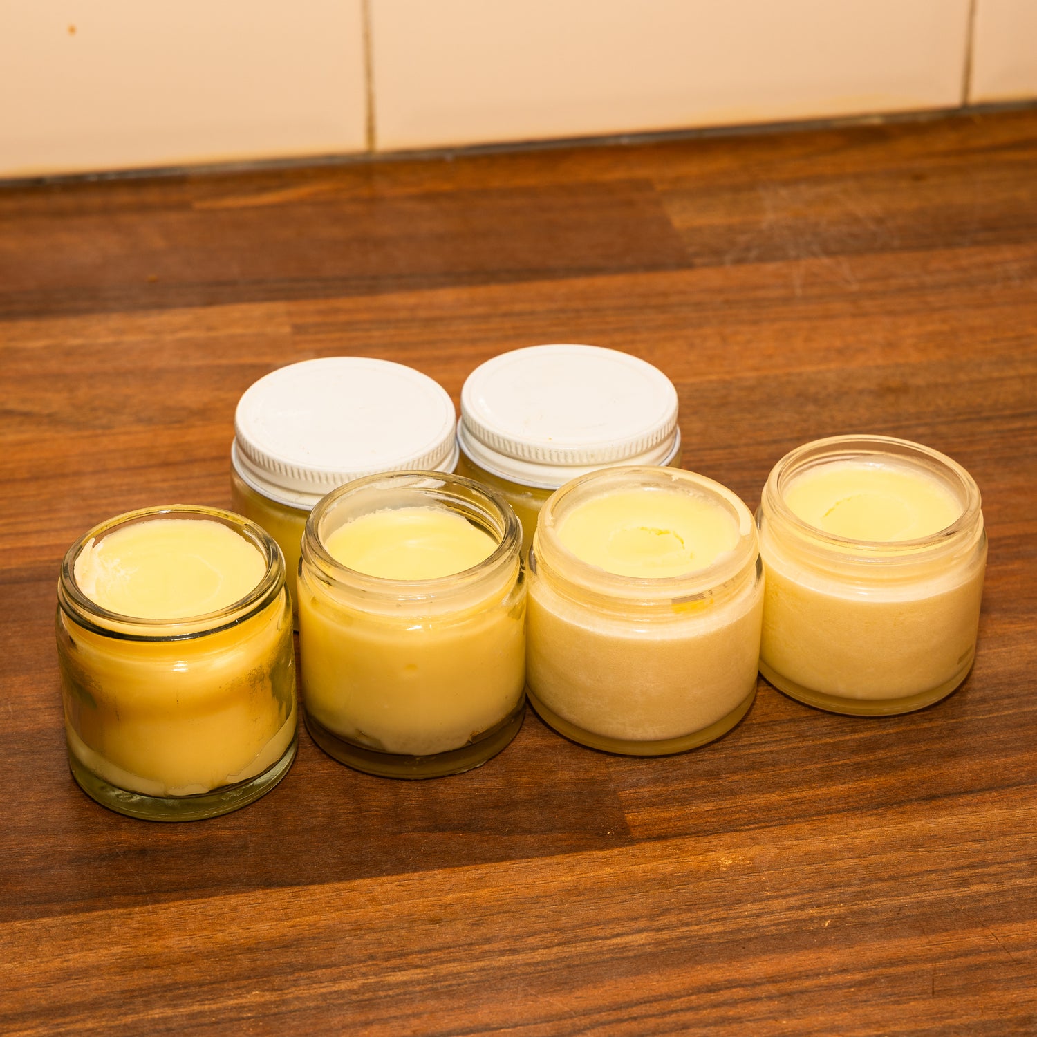 Five glass jars filled with tallow balm displayed on a wooden countertop. The jars in the front row are uncapped, revealing a variety of creamy textures and slight color differences, while three white plastic lids rest behind them. The setting is warmly lit, highlighting the artisanal, handcrafted nature of the balms.