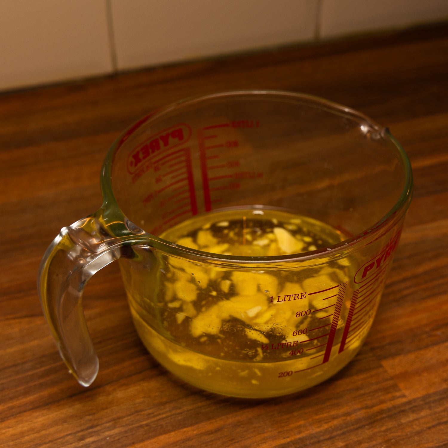 Glass Pyrex measuring jug filled with partially melted tallow and oils, placed on a wooden countertop. The mixture is in the process of blending, with visible chunks suspended in golden liquid. Measurement markings in red are printed on the jug.