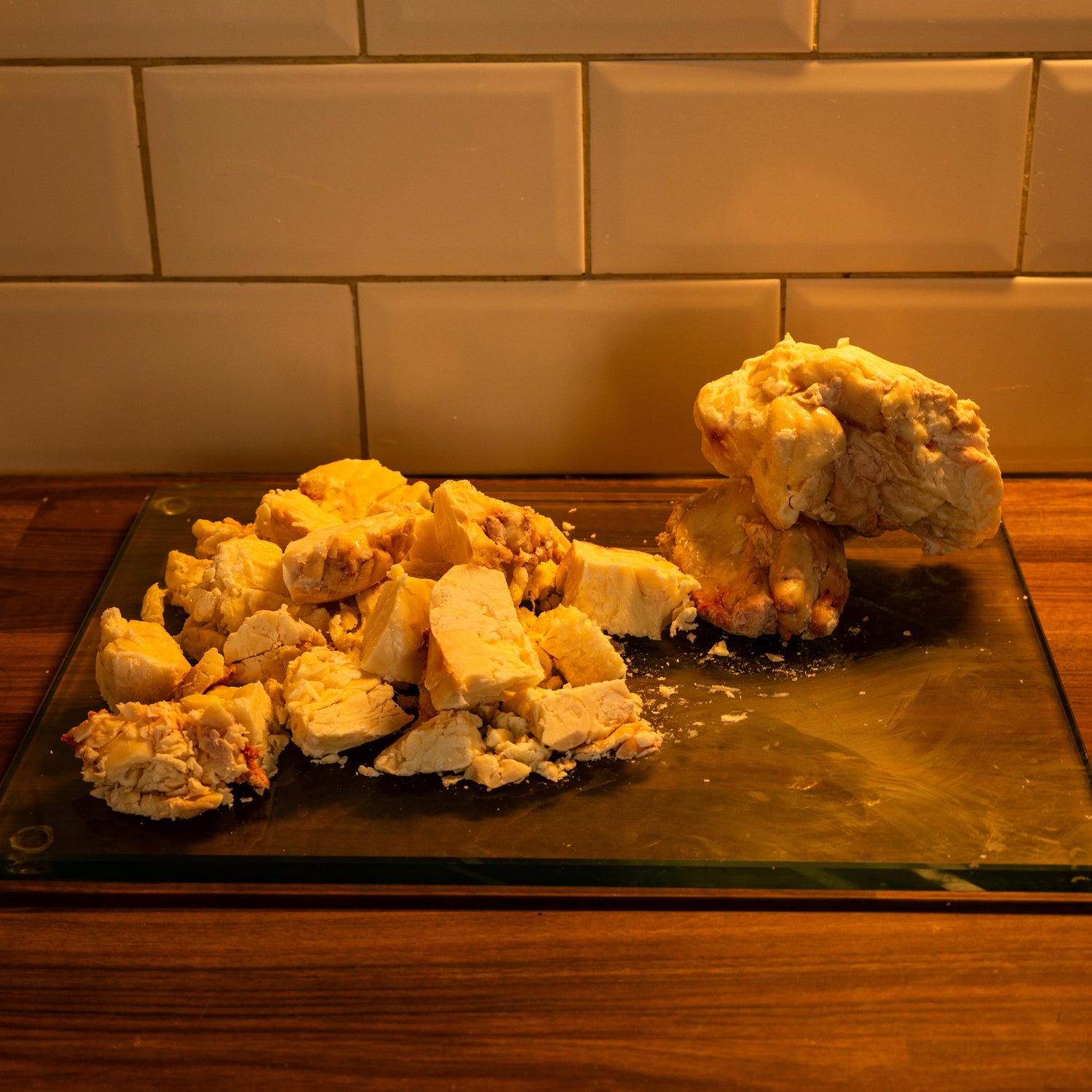 Raw chunks of beef suet placed on a glass cutting board on a wooden countertop, with white tiled walls in the background under warm indoor lighting.