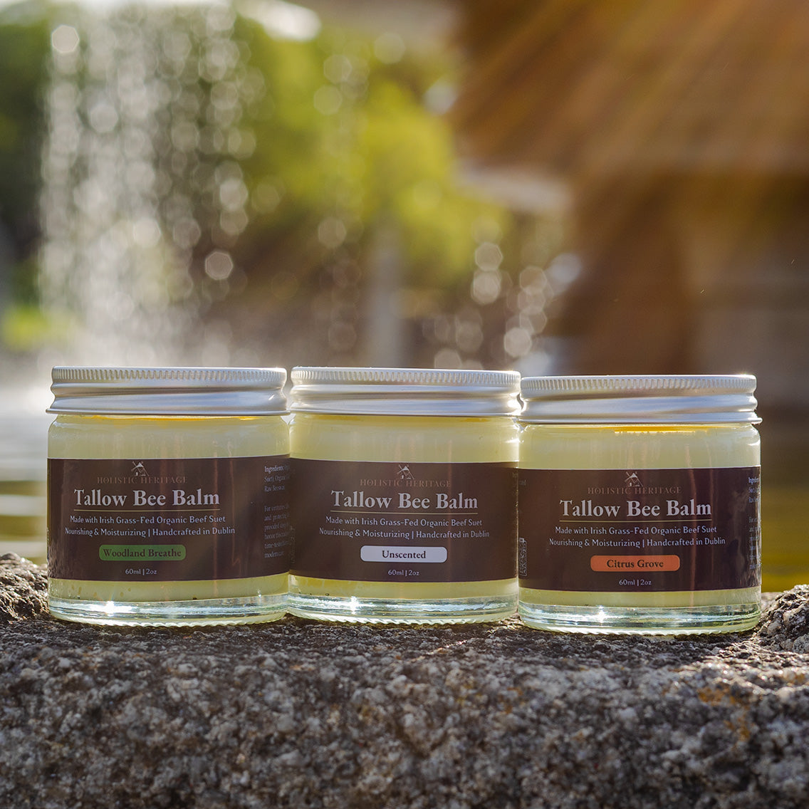 Three Holistic Heritage Tallow Bee Balm jars—Woodland Breathe, Unscented, and Citrus Grove—lined up on a stone surface with soft sunlight and a blurred fountain background.