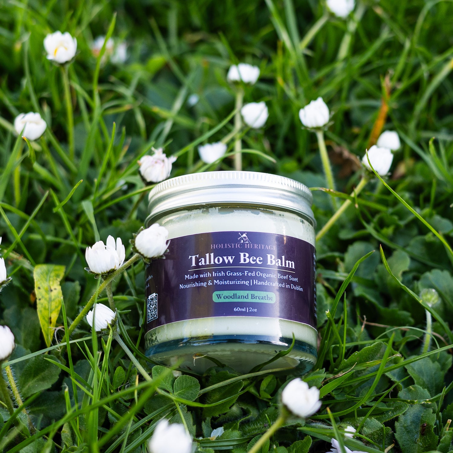 Glass jar of Holistic Heritage Tallow Bee Balm (Woodland Breathe scent) resting on green grass surrounded by small white flowers.